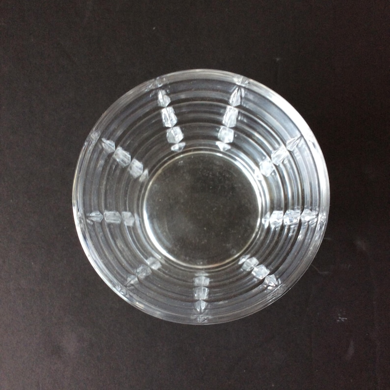 Op de afbeelding: Een heldere glazen kom met een cirkelvormig ontwerp met concentrische ringen en verticale ribbels die vanuit het midden naar de rand uitstralen. De kom heeft een gladde binnenkant en een getextureerde buitenkant met patronen.