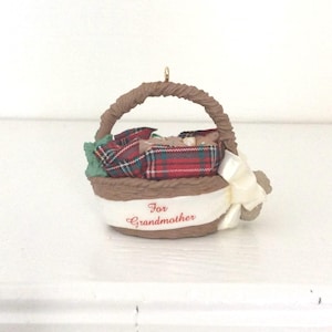 May include: A brown wicker basket ornament with a red and green plaid blanket inside. The basket has a white ribbon with the words "For Grandmother" written in red.