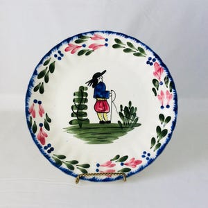 Puede incluir: Un plato de cerámica blanco con un borde floral azul, verde y rosa. El centro del plato presenta una ilustración pintada a mano de una persona con una chaqueta azul y pantalones rojos de pie en un campo verde.