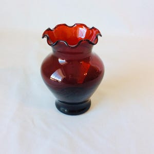May include: A deep red glass vase featuring a bulbous body and a wide, ruffled, crimped rim. The translucent glass has a smooth, polished finish with visible light reflections on its curved surface and a sturdy, circular base.