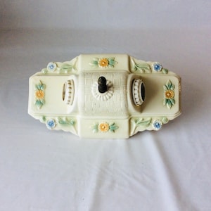 May include: A rectangular cream-colored glass ceiling light fixture featuring a central basket-weave texture, two light bulb sockets, and a dark metal finial. The edges are decorated with embossed floral motifs in yellow, blue, and green.