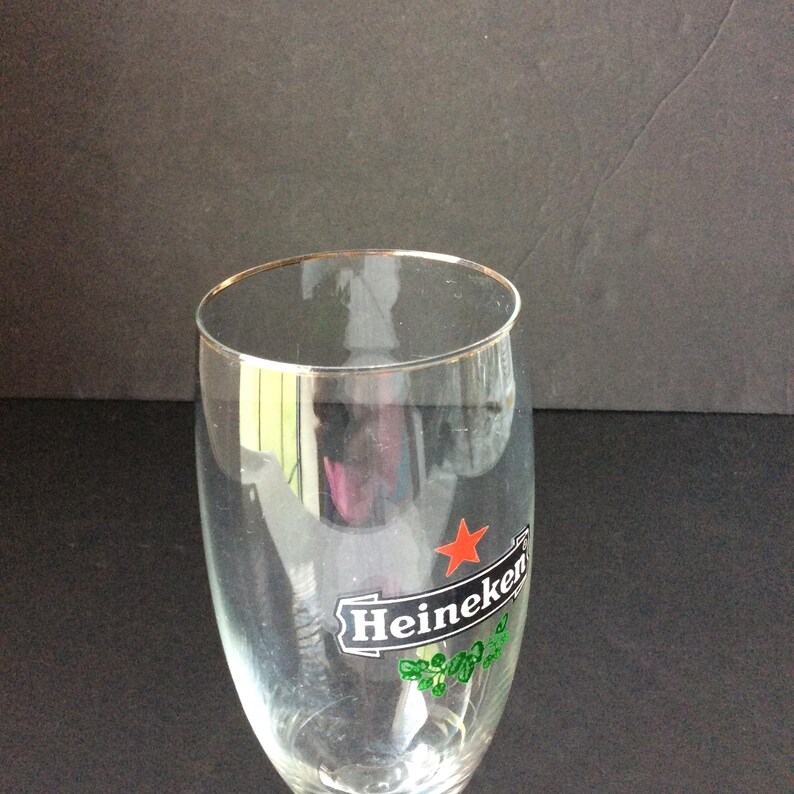May include: A clear beer glass with a gold rim. The glass features the Heineken logo, including a red star and green shamrocks. The glass is designed for beer.