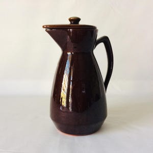 May include: A dark brown ceramic pitcher with a lid and a handle. The pitcher has a glossy finish and a slightly rounded shape.