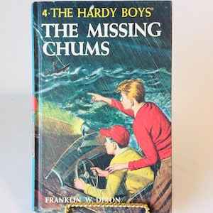 May include: Vintage hardcover book titled "The Hardy Boys: The Missing Chums." The cover art depicts two young men in a boat, with one pointing towards the stormy sea. The book is displayed on a gold-colored stand.