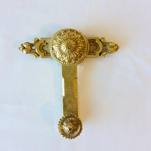 Puede incluir: Un tirador o manija de puerta vintage en tono latón con detalles florales y geométricos ornamentados. El tirador tiene forma de T con un diseño circular en la parte superior e inferior, y elementos decorativos en la barra horizontal.