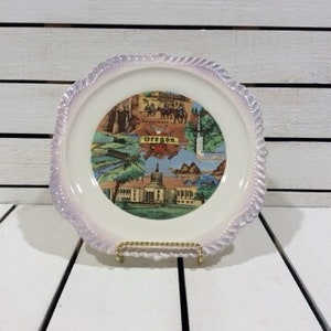 May include: A vintage decorative plate featuring a colorful collage of Oregon landmarks and scenes. The plate has a white center with a detailed image and a light purple, scalloped edge. It is displayed on a gold-colored metal stand.