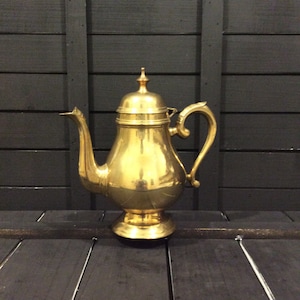 May include: A polished brass teapot featuring a rounded body, a pedestal base, a curved handle, and a long, slender spout. The lid is topped with a small, pointed finial. The metal surface has a reflective, golden finish with visible highlights.