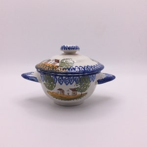 May include: A ceramic lidded bowl with two side handles, featuring a hand-painted landscape scene of houses and trees in green, brown, and yellow. The rim and lid handle are decorated with a blue patterned border on a white background.