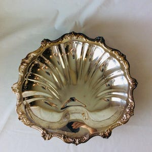 May include: A silver-colored, shell-shaped serving dish with a decorative, ornate rim. The dish has a fluted interior and a reflective surface, suggesting it is made of metal. The rim has a detailed, embossed design.