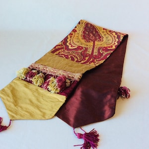 May include: A decorative table runner with a paisley design in gold and burgundy. The runner has gold and burgundy tassels at each end and a decorative trim with tassels in the middle. The runner is made of a silky material.