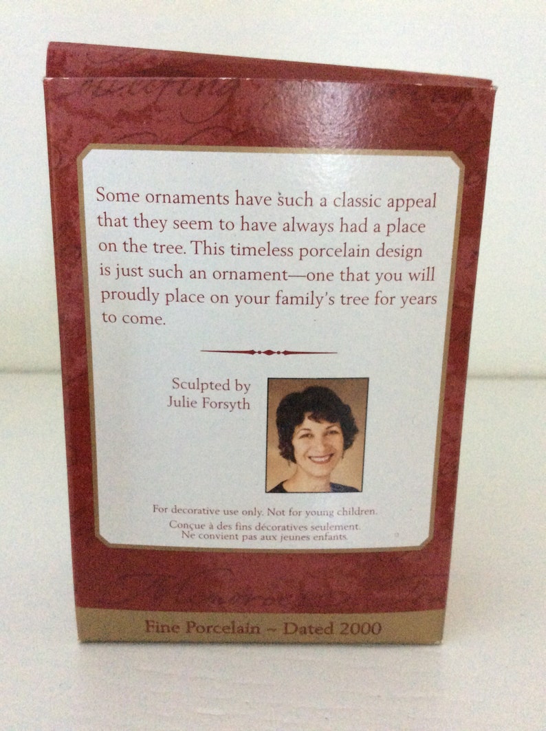 May include: A maroon box with text about a porcelain ornament. The text reads "Some ornaments have such a classic appeal...Sculpted by Julie Forsyth...Fine Porcelain - Dated 2000". A photo of Julie Forsyth is included.