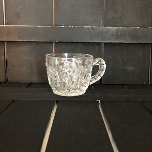 May include: A clear, cut-glass teacup with a handle. The cup features a detailed, repeating pattern of diamond and star shapes. The handle is also textured. The cup is sitting on a black surface.