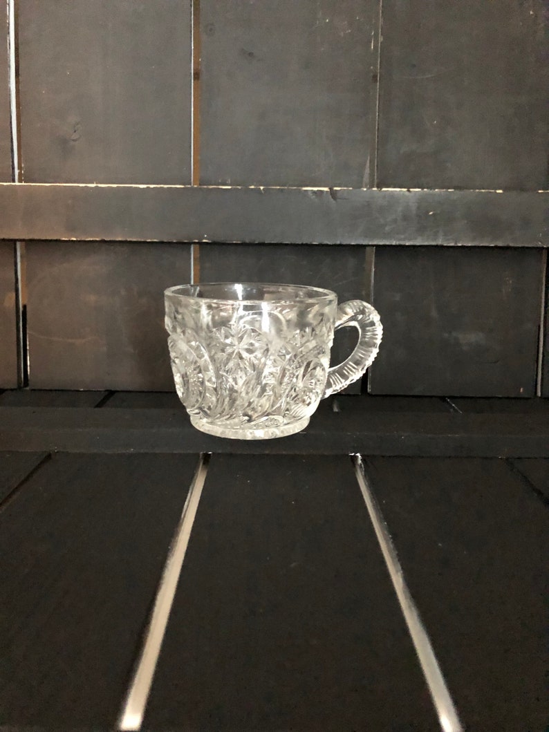 May include: A clear, cut-glass teacup with a handle. The cup features a detailed, repeating pattern of diamond and star shapes. The handle is also textured. The cup is sitting on a black surface.