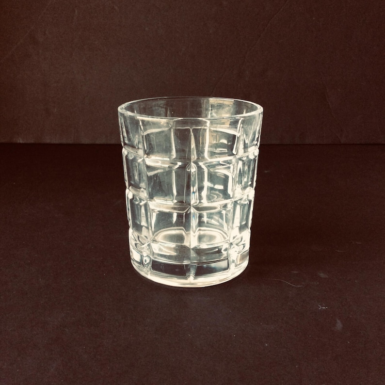 Op de afbeelding: Een helder glazen drinkglas met een herhalend rasterpatroon van verhoogde vierkante facetten over het buitenoppervlak. De beker heeft een cilindrische vorm met een dikke bodem en een gladde rand, ontworpen voor het serveren van diverse dranken.