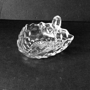 May include: Clear glass dish with a faceted, geometric design. The dish has a leaf-like shape with a handle. The glass is transparent, allowing light to pass through, and sits on a dark surface.