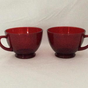 Könnte beinhalten: Zwei rubinrote Teetassen aus Glas mit Henkeln. Die Tassen haben eine abgerundete Schalenform und stehen auf einem kleinen Sockel. Die Tassen bestehen aus einem durchscheinenden Glasmaterial.