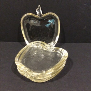 Puede incluir: Juego de platos de vidrio transparente en forma de manzana. El juego incluye una tapa y varios platos a juego. Los platos están apilados y la tapa se coloca encima. El vidrio tiene un ligero tinte amarillo.