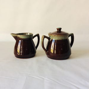 May include: A brown ceramic creamer and sugar bowl set. The creamer has a spout and handle, while the sugar bowl has a lid and handles. Both pieces have a green band near the top and are on a white background.