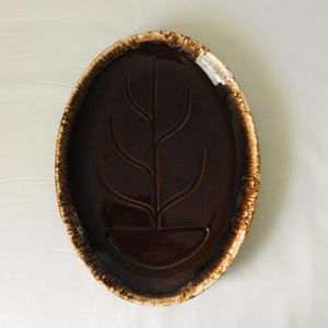 May include: An oval-shaped ceramic platter featuring a deep, glossy dark brown center with a debossed stylized plant motif. The rim is finished with a textured, mottled light brown and cream glaze that creates a distinct contrast to the dark interior.