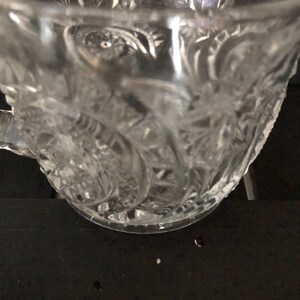 May include: Clear glass teacup with ornate floral and starburst patterns. The cup has a handle and a wide rim. The glass is transparent, allowing light to pass through, and is set against a dark background.