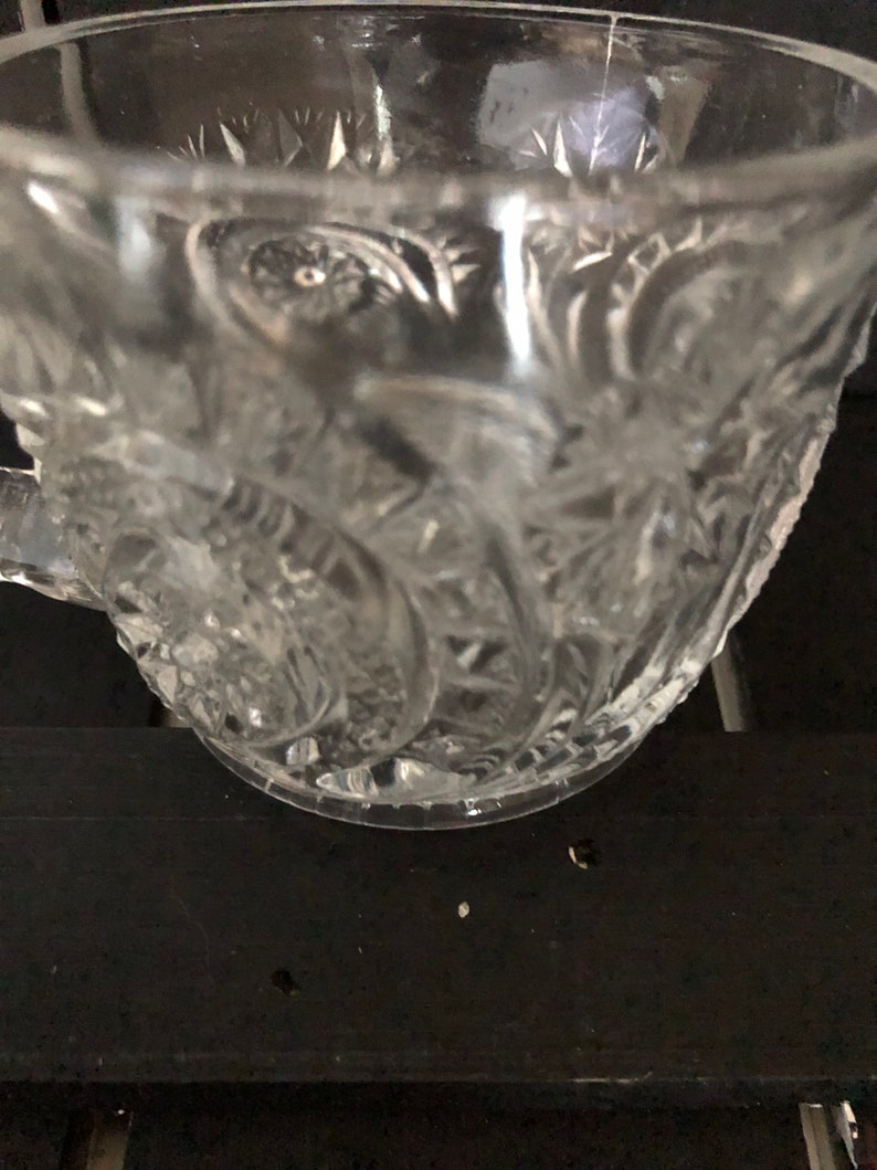 May include: Clear glass teacup with ornate floral and starburst patterns. The cup has a handle and a wide rim. The glass is transparent, allowing light to pass through, and is set against a dark background.