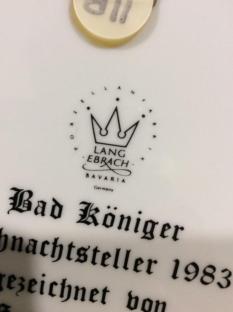 以下が含まれることがあります： 白磁のプレートに、黒と白のロゴが描かれており、「Lang Ebrach Bavaria Germany」と王冠が書かれています。 「Bad K&ouml;niger Weihnachtsteller 1983」というテキストが黒で書かれています。