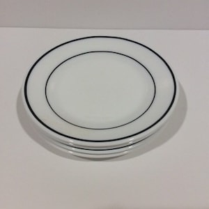 May include: A stack of white ceramic plates with a black border. The plates are round and have a simple, elegant design. The plates are stacked on top of each other.