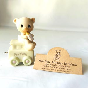 May include: A porcelain figurine of a bear sitting on a toy train. The bear is holding a bottle and the train says "For Baby". The figurine is part of a series called "May Your Birthday Be Warm" and is one of eight collectibles in the Birthday Series Train. A Precious Moments Porcelain Figurine.