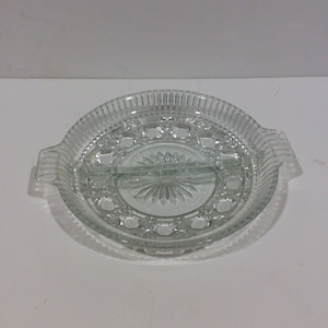 Op de afbeelding: Transparante glazen serveerschaal met twee secties en een centraal ontwerp. De schaal heeft een cirkelvorm met een geribbelde buitenrand en twee handvatten. Het midden heeft een sterpatroon. Een decoratief stuk voor het serveren van snacks of hapjes.
