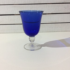 May include: A cobalt blue glass goblet with a clear glass stem and base. The goblet is sitting on a white surface.
