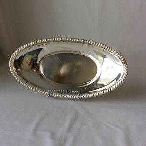 May include: An oval silver-toned serving tray with a polished, reflective surface and a decorative rope-style border along the rim. The tray is displayed upright on a small, thin metal stand, showcasing its smooth, metallic finish and oval shape.