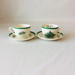 Puede incluir: Dos tazas de té de cerámica color crema con platillos a juego y borde verde. Una taza presenta un diseño de rama de acebo, mientras que la otra muestra un árbol de Navidad decorado. Ambos platillos están decorados con motivos de hojas de acebo.