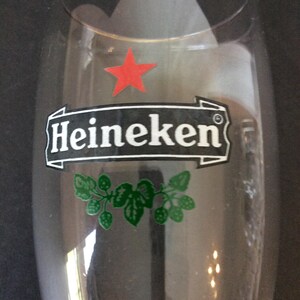 May include: A clear beer glass with the Heineken logo. The logo features the word "Heineken" in white on a black banner, a red star, and green leaf and hop illustrations.
