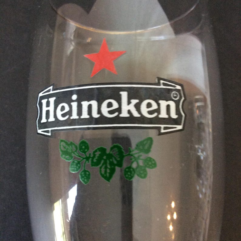 May include: A clear beer glass with the Heineken logo. The logo features the word "Heineken" in white on a black banner, a red star, and green leaf and hop illustrations.