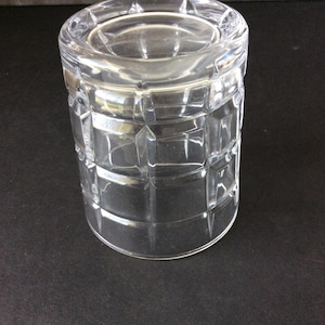 Op de afbeelding: Een helder glazen drinkglas met een geometrisch rasterpatroon van verhoogde vierkanten over het buitenoppervlak. Het vat heeft een cilindrische vorm met een dikke, gladde rand en een platte bodem, wat een transparant ontwerp met lichtreflecties toont.
