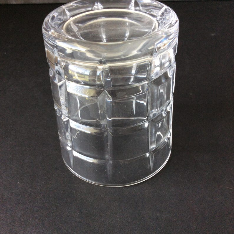Op de afbeelding: Een helder glazen drinkglas met een geometrisch rasterpatroon van verhoogde vierkanten over het buitenoppervlak. Het vat heeft een cilindrische vorm met een dikke, gladde rand en een platte bodem, wat een transparant ontwerp met lichtreflecties toont.