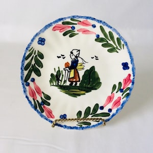May include: A round ceramic plate with a scalloped edge featuring a blue painted rim. The center displays a hand-painted illustration of a figure in a bonnet and apron standing in a field. The border is decorated with pink flowers and green leaves.