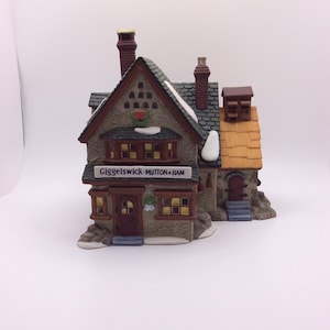Puede incluir: Una detallada miniatura de cerámica de un edificio, posiblemente una tienda o pub, con el letrero "Giggelswick-MUTTON & HAM". El edificio tiene un tejado marrón, chimeneas de ladrillo y detalles de nieve. Las ventanas tienen un brillo cálido.