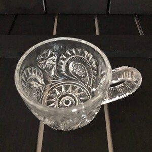 May include: Clear glass mug with intricate, embossed floral and geometric patterns. The handle is also textured. The mug is sitting on a black surface.