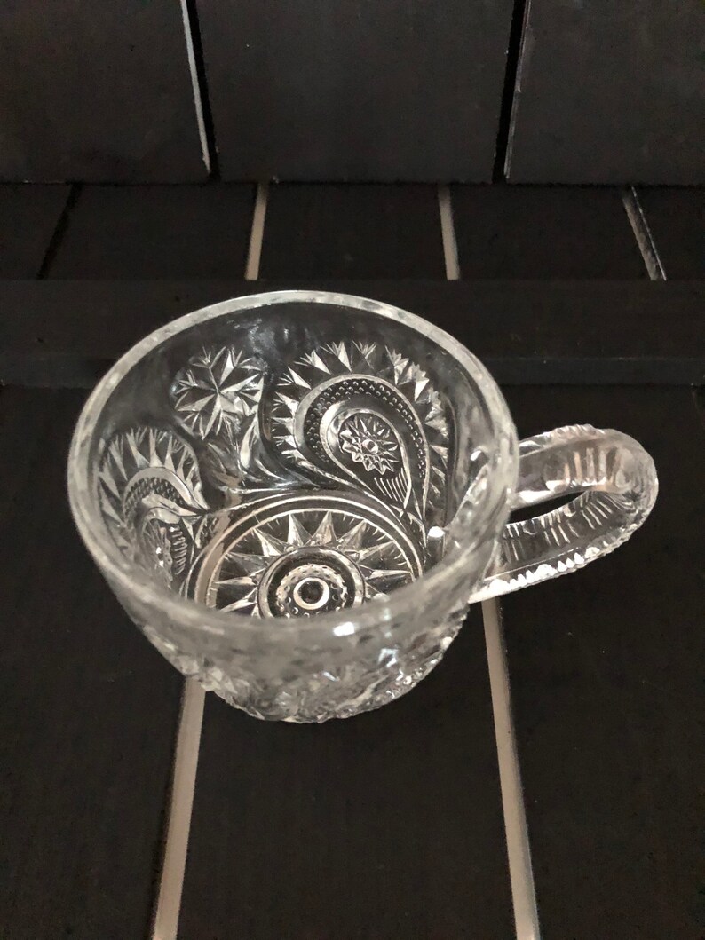 May include: Clear glass mug with intricate, embossed floral and geometric patterns. The handle is also textured. The mug is sitting on a black surface.