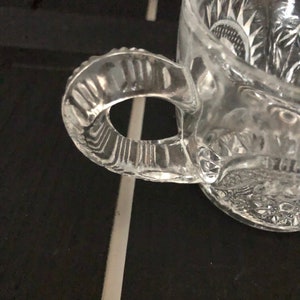 May include: A clear glass mug with a decorative handle and etched floral patterns. The mug is made of clear glass and has a textured handle. The handle is attached to the side of the mug. The mug is sitting on a dark surface.