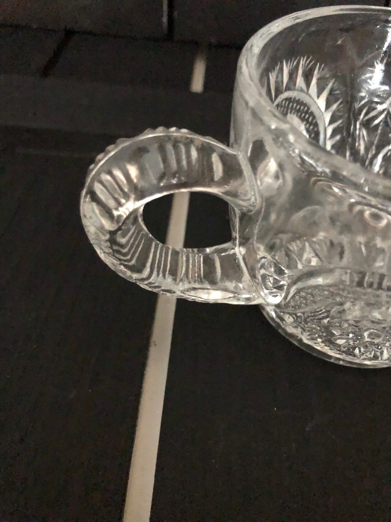 May include: A clear glass mug with a decorative handle and etched floral patterns. The mug is made of clear glass and has a textured handle. The handle is attached to the side of the mug. The mug is sitting on a dark surface.