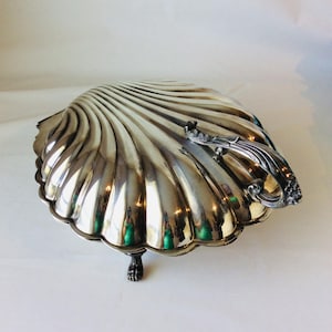 以下が含まれることがあります： 磨き上げられた金属仕上げの大きなホタテ貝の形をした銀色の盛り付け皿。この皿は片側に装飾的で華やかなハンドルを備えており、貝殻構造の底にある3つの小さな詳細な脚によって支えられています。