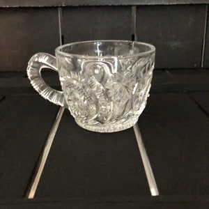 May include: A clear glass teacup with a handle. The cup features a detailed, embossed floral pattern around the exterior. The handle is also textured. The teacup is sitting on a dark surface.