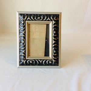 May include: A rectangular picture frame featuring a thick, ornate border with a black finish and silver-toned highlights. The frame has a smooth silver inner edge and a smooth silver outer edge, surrounding a rectangular opening with a beige insert.