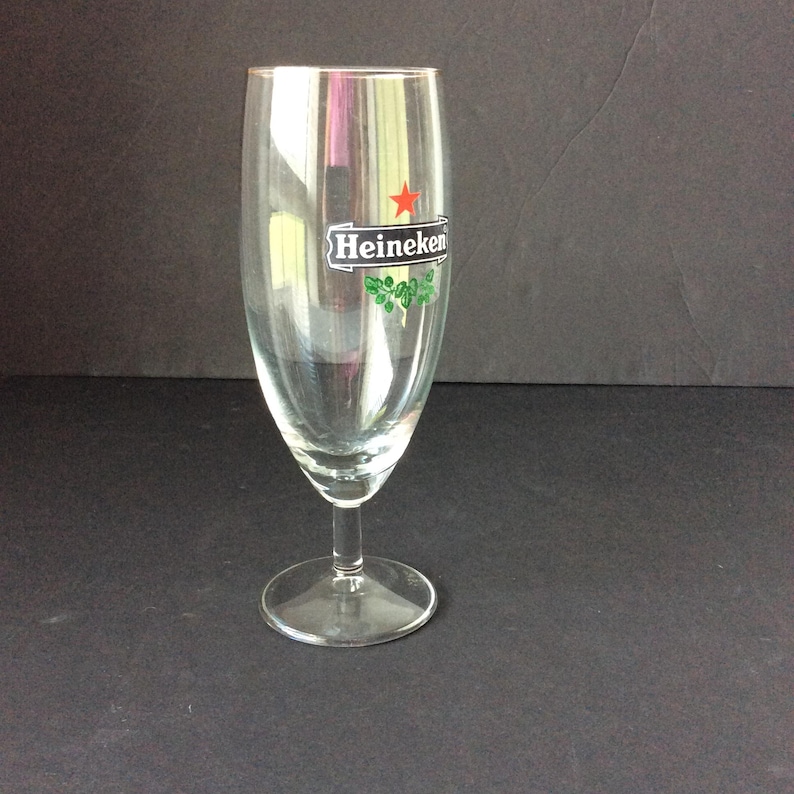 May include: A clear, stemmed beer glass with the Heineken logo, featuring a red star and green accents. The glass has a tall, slender bowl and a round base. The glass is empty and sits on a dark surface.