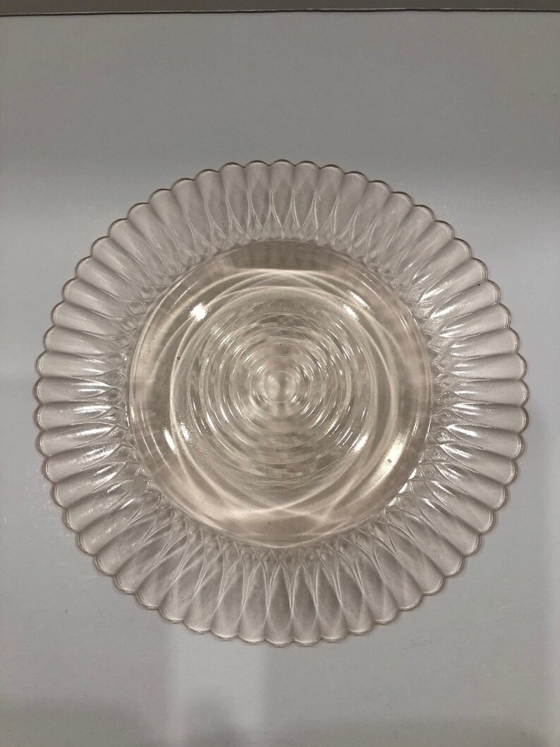 Puede incluir: Plato de vidrio transparente con borde festoneado. El centro del plato presenta c&iacute;rculos conc&eacute;ntricos. El plato es transparente y probablemente se utiliza para servir alimentos.