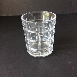Op de afbeelding: Een helder glazen drinkglas met een geometrisch rasterpatroon van verhoogde vierkanten over het buitenoppervlak. De beker heeft een cilindrische vorm met een dikke bodem en een gladde rand, wat zorgt voor een transparant ontwerp.