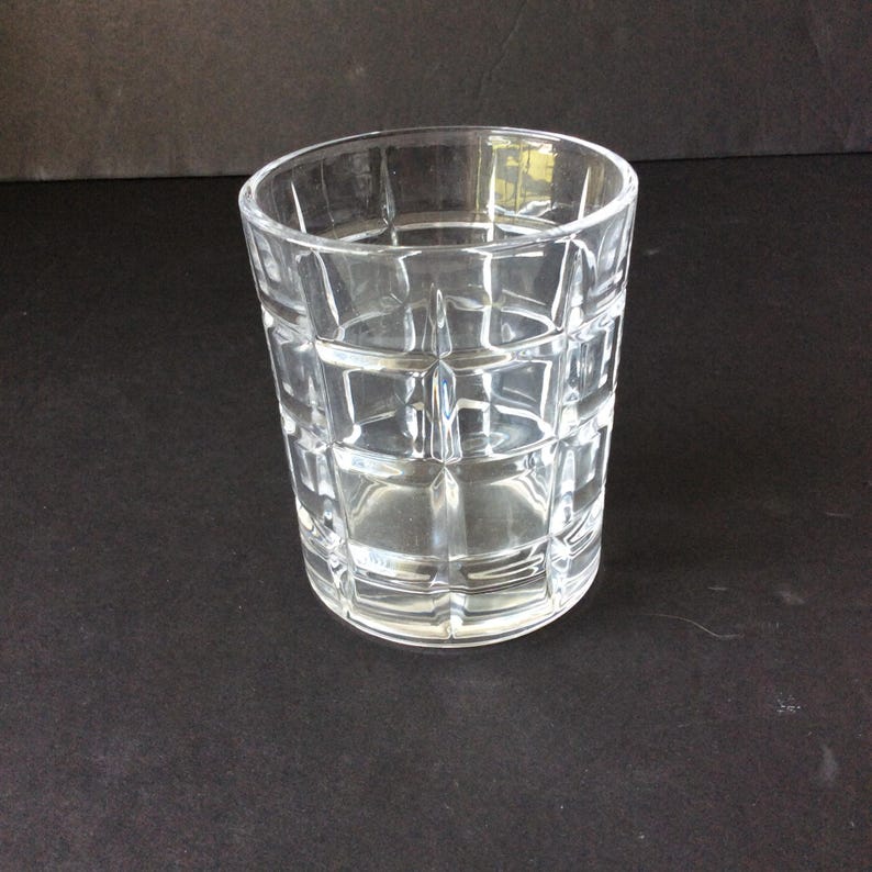 Op de afbeelding: Een helder glazen drinkglas met een geometrisch rasterpatroon van verhoogde vierkanten over het buitenoppervlak. De beker heeft een cilindrische vorm met een dikke bodem en een gladde rand, wat zorgt voor een transparant ontwerp.