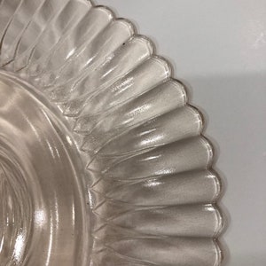 Puede incluir: Primer plano de un plato de vidrio transparente con borde festoneado. El plato presenta un dise&ntilde;o texturizado y acanalado que se extiende desde el centro hasta el borde. El vidrio transparente permite el paso de la luz, sobre un fondo blanco.